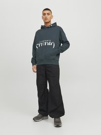 Mørkegrøn hættetrøje med hvid "originals STUDIO" tekst, kengurulomme, ribbet kant, parret med løse sorte bukser og grå sneakers.