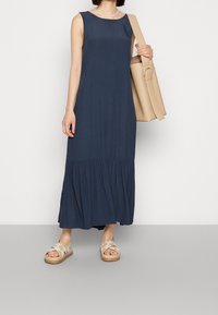 Sininen hihaton maksimekko, jossa on röyhelöhelma, yhdistettynä beigeihin ristikkäin nauhoihin sekä suureen beigeen olkalaukkuun.