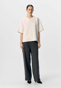 Överdimensionerad ljusrosa sweatshirt med korta ärmar, parat med grå vida byxor. Modell som bär svarta spetsiga skor, stående mot en neutral bakgrund.