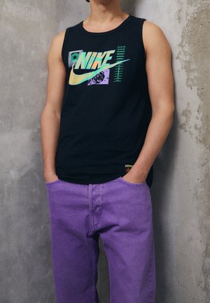 Osoba nosząca czarny top Nike z wielokolorowym logo i fioletowe spodnie, stojąca na tle teksturowanej szarej betonowej ściany.