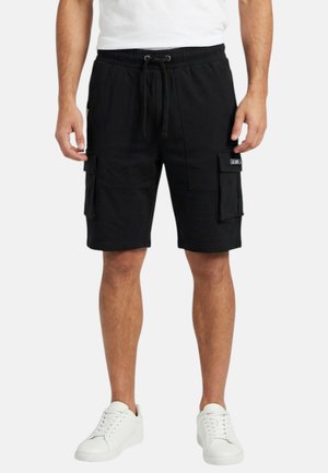 Homme portant un short cargo noir avec taille ajustable par cordon, assorti de baskets blanches et d'un t-shirt blanc, debout devant un fond uni.