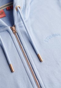 Sudadera con capucha azul claro con cremallera, confeccionada en tela suave, que presenta una cremallera dorada, detalles de marca y un sutil logo bordado.