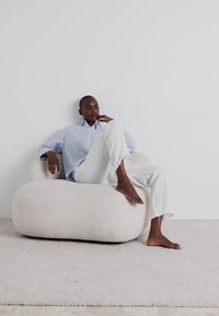 Kurvig, off-white klädd stol med mjukt, texturerat tyg. En person i lösa, ljusa kläder sitter avslappnat och sträcker ut ett ben.