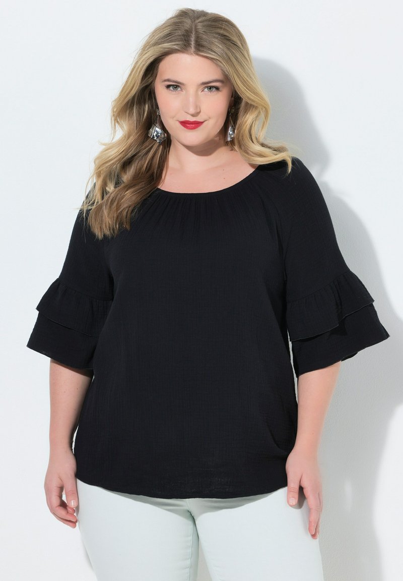 Blonde vrouw met golvend haar, rode lippenstift, zilveren oorbellen, een top met zwart gerafelde mouwen en een witte broek tegen een effen witte achtergrond.