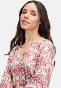Bunt gemusterte Bluse mit einem geometrischen Design in pinken, gelben und braunen Akzenten. Hergestellt aus leichtem Stoff mit einem V-Ausschnitt.
