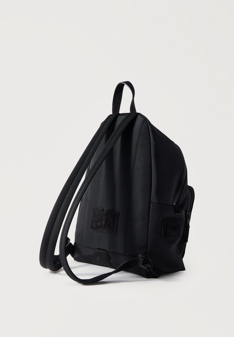 Sac à dos compact noir avec deux bretelles ajustables et fermeture éclair, debout sur un fond clair uni.