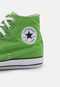 Sneaker cano alto verde em lona com atacadores brancos, patch de logotipo preto e branco, e sola de borracha branca apresentando riscas pretas.