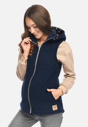 Lächelnde Frau in einem beigen Sweatshirt und einer navyblauen, ärmellosen Weste mit Reißverschluss und sternenbedruckter Kapuze, die mit einer Hand in der Tasche posiert.
