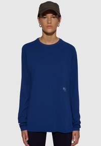 Długi rękaw, teksturowana niebieska koszulka sportowa z rękawami raglanowymi i małym logo po lewej stronie z przodu. Materiał wydaje się oddychający i lekki.