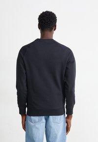 Homme aux cheveux courts et bouclés portant un sweat-shirt noir et un jean bleu clair, debout, dos tourné, sur un fond blanc uni.
