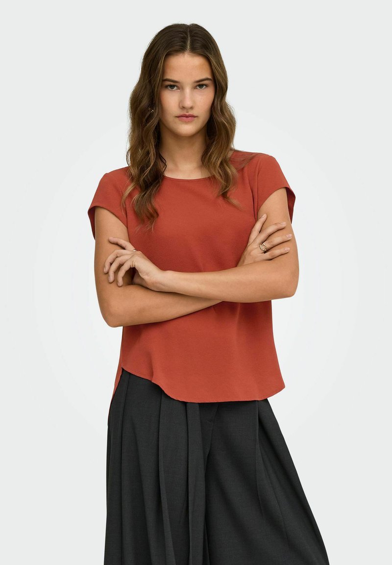 Femme aux longs cheveux ondulés portant un haut à manches courtes couleur rouille et un pantalon noir ample, debout les bras croisés sur un fond uni.