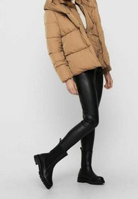 Giacca imbottita color fango con chiusura frontale con cerniera, indossata sopra leggings in ecopelle nera e stivali neri alla caviglia con suole testurizzate e cerniere laterali.