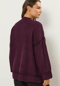 Pull en maille de couleur violet profond avec une finition texturée. Présente des accents de couture noire le long des manches et de l'ourlet. Design à coupe décontractée.