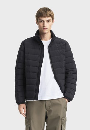 Jeune homme portant une veste matelassée noire sur un t-shirt blanc et un pantalon cargo kaki, debout devant un fond uni.