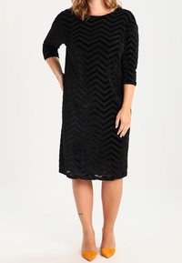 Black knee-length dress with a chevron pattern, made of textured fabric. Features three-quarter sleeves and a round neckline, paired with yellow heels.