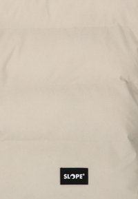 Beige pufferjacka med horisontellt quiltade sektioner, slät textur och en svart logotypapplikation märkt "SLOPE" nederst.