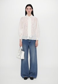 Chemisier blanc translucide avec motif floral, col noué et poignets boutonnés, associé à un jean bleu à jambes larges et un petit sac à main blanc.