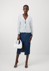 Λευκή μπλούζα με κουμπί γαζί και v-λαιμόκοψη, συνδυασμένη με τζιν φούστα με μπροστινή σχισμή, κρατώντας μια καπιτονέ άσπρη τσάντα.