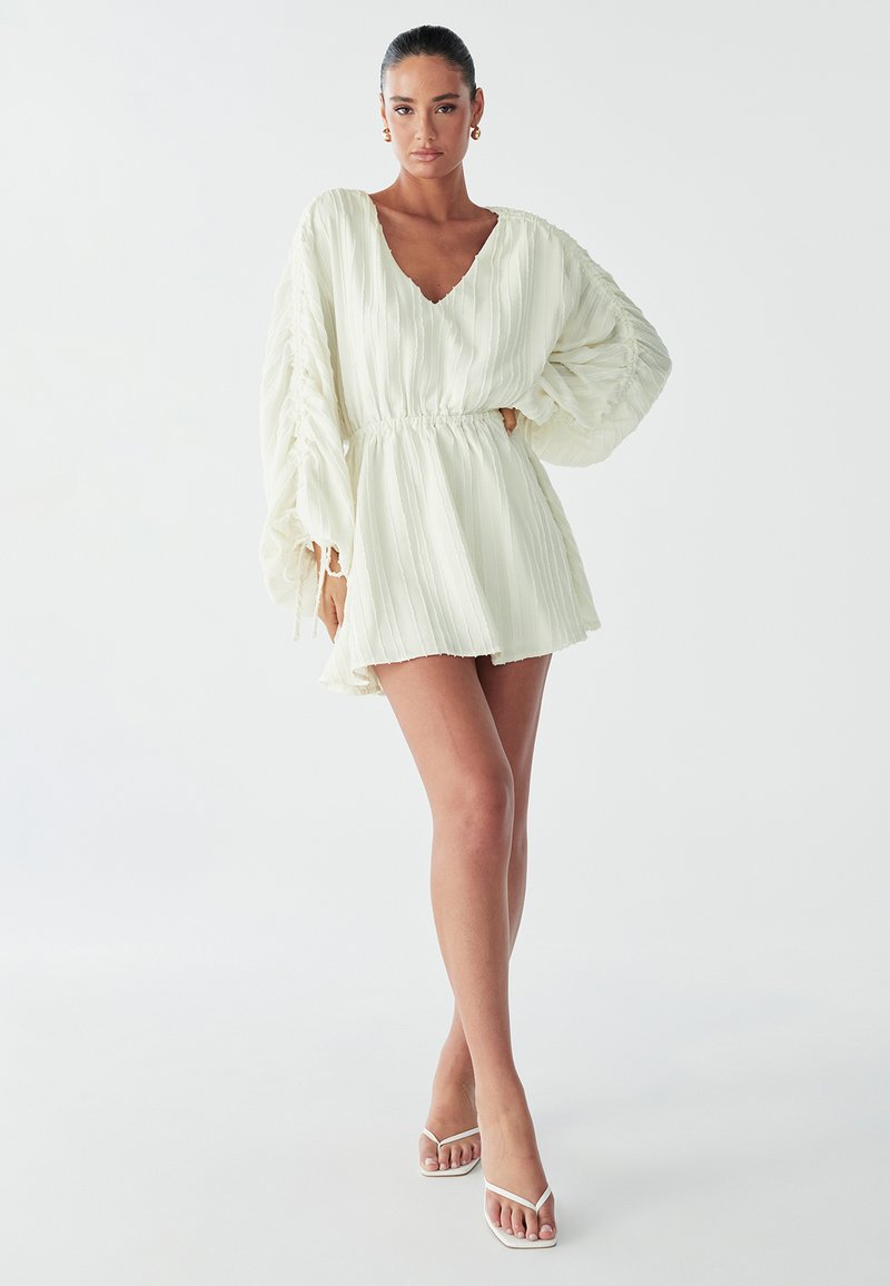 Model poseert zelfverzekerd in een lichte, geplooide jurk met voluminieuze mouwen, gecombineerd met minimalistische witte sandalen tegen een simpele achtergrond.