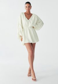 Model poseert zelfverzekerd in een lichte, geplooide jurk met voluminieuze mouwen, gecombineerd met minimalistische witte sandalen tegen een simpele achtergrond.