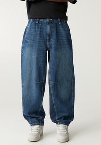 Jean en denim bleu clair avec une coupe décontractée, comportant des poches avant, des coutures visibles et une texture légèrement décolorée. Associé à des baskets blanches.