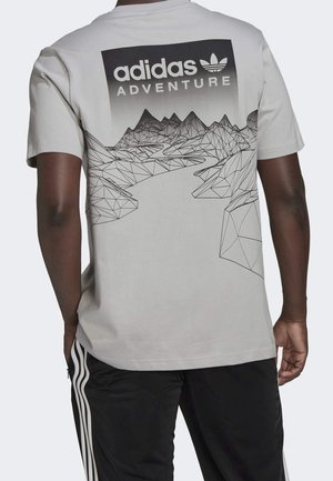 Homme portant un t-shirt Adidas gris clair avec un motif géométrique noir de montagne et de rivière ainsi que le texte « adidas ADVENTURE » au dos.