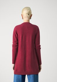 Röd långärmad cardigan med en mjuk textur, ribbad nederkant och manschetter, med en klassisk rund halsringning och förlängd längd.