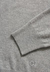 Grauer Strickpullover mit geripptem Bund und kleinem gestickten "OJ"-Logo. Glatte Textur mit einem dezenten Muster im Stoff.