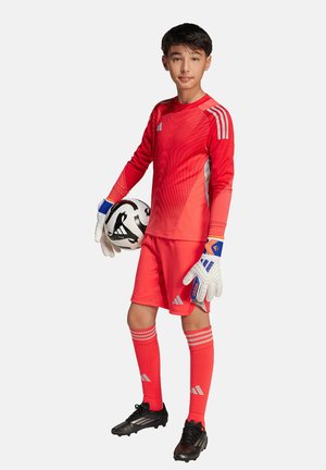Uniforme de gardien de but rouge en matériau synthétique, avec des accents rayés, manches longues, shorts assortis et gants. Ballon de football noir en main.