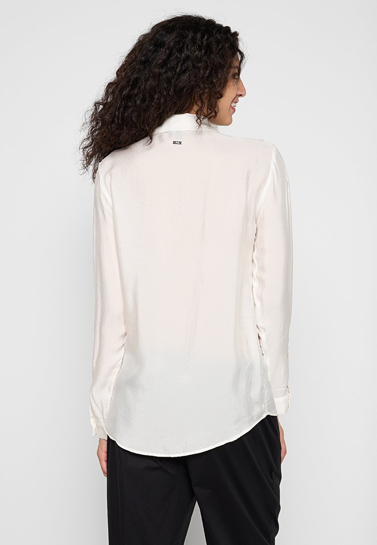 Blusa blanca de manga larga con un acabado texturizado, que presenta un dobladillo ligeramente curvado y un pequeño detalle de logo en el cuello trasero.