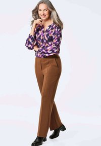 Paarse en roze gebloemde blouse, bruine op maat gemaakte broeken en zwarte haklaarzen. Het model heeft lang, golvend grijs haar en staat in een nadenkende pose.