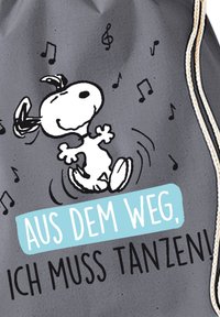 Un tessuto grigio presenta un cane cartone animato che balla, con corpo bianco e dettagli neri. Il testo in blu e nero recita "AUS DEM WEG, ICH MUSS TANZEN!"