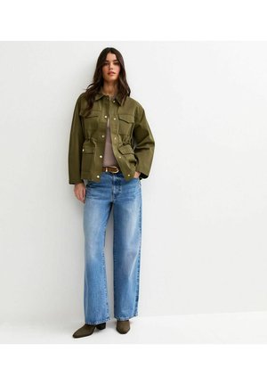 Kobieta ubrana w oliwkowo-zieloną zapinaną na guziki kurtkę z czterema kieszeniami, jasnobrązową bluzkę, szerokie niebieskie dżinsy i brązowe kozaki na obcasie, stoi opierając się o ścianę.