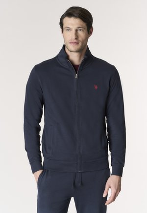 Uomo che indossa una giacca blu navy con zip e logo rosso sul petto, in piedi con le mani nelle tasche davanti a uno sfondo chiaro e semplice.