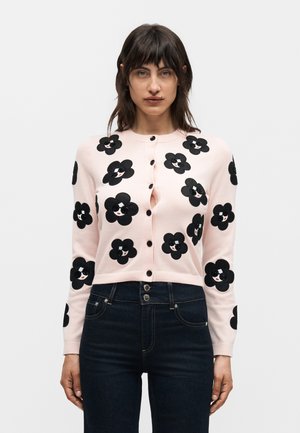 Kvinde iført en lyserød cardigan med knapper og sorte blomster med smilende ansigter, kombineret med højtaljede mørke jeans.