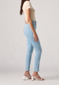 Vaqueros pitillo de talle alto en azul claro con un diseño clásico de cinco bolsillos y una placa de cuero con la marca en la parte trasera. Combinados con sandalias blancas de tacón cuadrado.