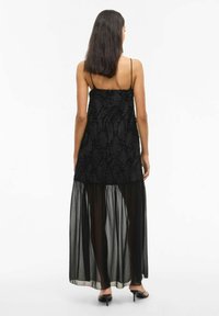 Juodas suknelė su tekstūruotu viršumi, kuriame yra iškilūs, kreivi raštai, ir skaidrus, plačios apimties sijonas. Ploni spaghetti tipo petnešėlės ir kulkšnis siekianti dizainas.