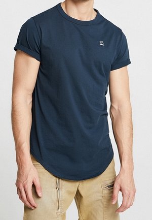 Homme portant un t-shirt bleu marine à manches retroussées et un pantalon cargo beige, se tenant contre un fond léger uni.