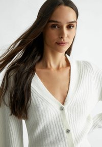 Cardigan bianco a coste con scollatura a V e maniche lunghe, caratterizzato da accenti decorativi in argento sui bottoni. Texture morbida e design aperto.