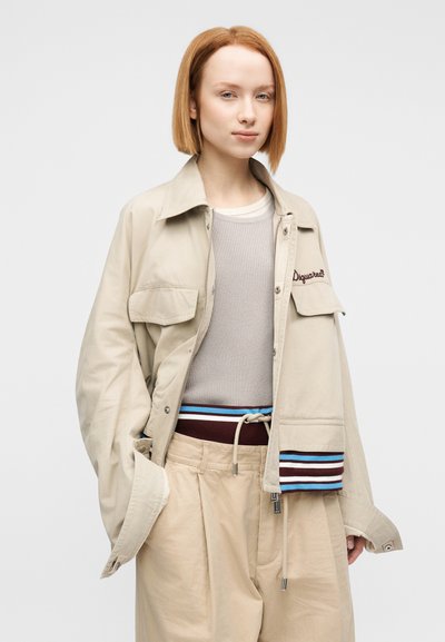 Μπεζ σακάκι από καμβά με φερμουάρ, δύο τσέπες στο στήθος, κεντημένο λογότυπο, φορεμένο πάνω από γκρι ριμπ τοπ και μπεζ πλισέ παντελόνι με ριγέ ζώνη.