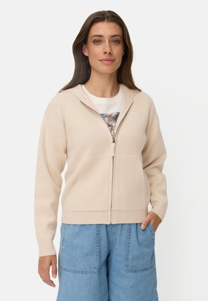 Vrouw met lang bruin haar die een beige hoodie met rits draagt, een wit T-shirt met print en blauwe wijde jeans met handen in de zakken.