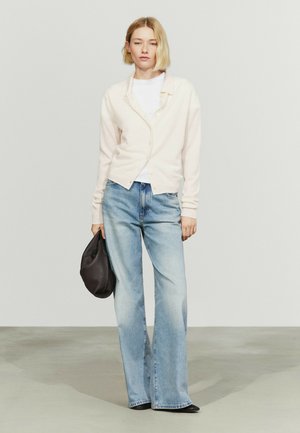Jeune femme aux cheveux blonds courts portant un cardigan blanc, une chemise blanche, un jean bleu clair, tenant un sac noir, debout sur un sol gris avec un fond clair uni.