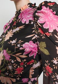 Černé květinové šaty s jasně růžovými, oranžovými a zelenými květinami. Rybkový výstřih a nabírané rukávy dodávají designu objem.