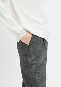 Osoba w białej bluzie z długim rękawem, z ręką w kieszeni szarych spodni w deseń herringbone, na jednolitym tle.