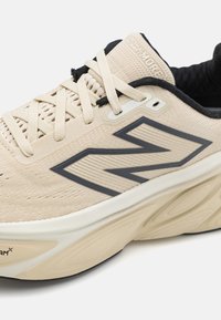 Baskets de sport beige et noir avec une semelle épaisse, logo "N" noir sur le côté, lacets beiges et col rembourré noir sur fond blanc.