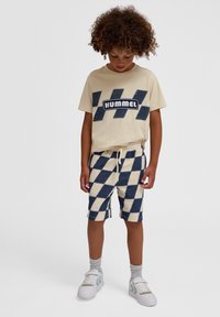 Krämfärgad t-shirt med marinblå logotyp och chevronm önster; rutiga beige och marinblå shorts. Vita sneakers med detaljer och grå strumpor.