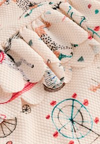 Textiles mit einer hellrosa Hintergrundfarbe, die ein verspieltes Muster aus Fahrrädern, bunten Formen und gewellten Kanten aufweisen.