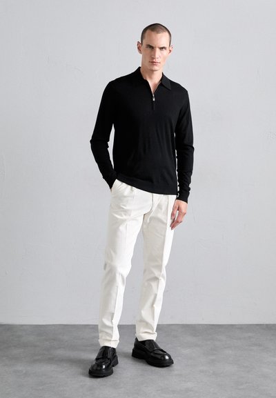 Homem vestido com camisa preta de mangas longas e gola com fecho, calças brancas e sapatos mocassim pretos e grossos, a ficar em pé sobre um chão cinza, contra uma parede cinza-clara.