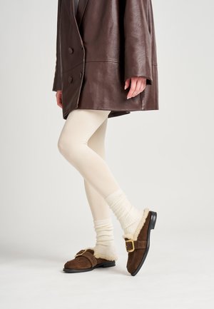 Des chaussures à enfiler en daim marron avec une doublure crémeuse et moelleuse et une boucle dorée, associées à des leggings crémeux et des chaussettes côtelées crémeuses, sous un manteau en cuir marron.
