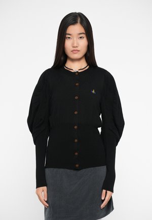 Cardigan nero a costine con maniche a sbuffo, dotato di bottoni marroni e un piccolo design ricamato. Indossato sopra una gonna grigia.